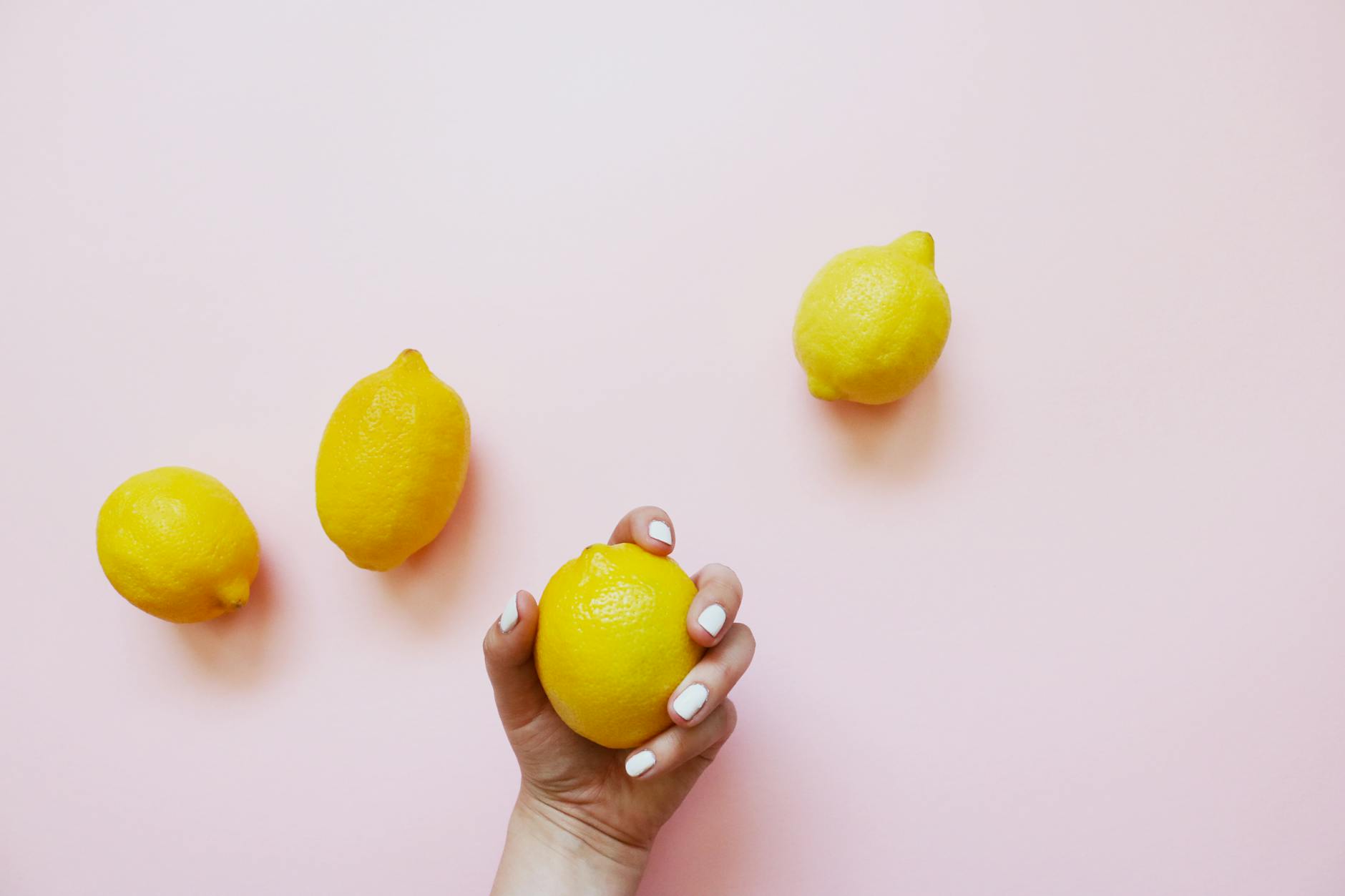 A hand holding a fresh lemon on a soft pink background with additional lemons surrounding it